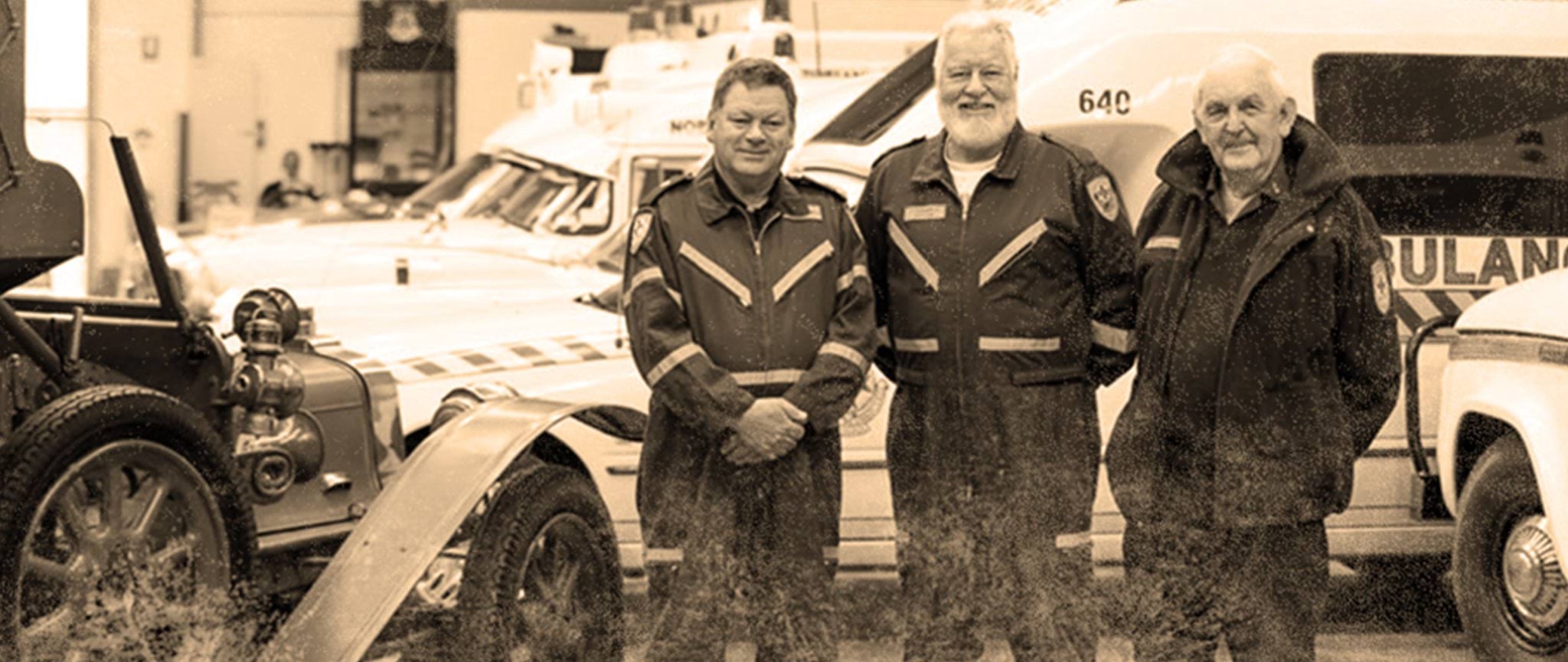 Three retired paramedics and first responders standing in front of several vintage ambulance vehicles parked inside a garage.