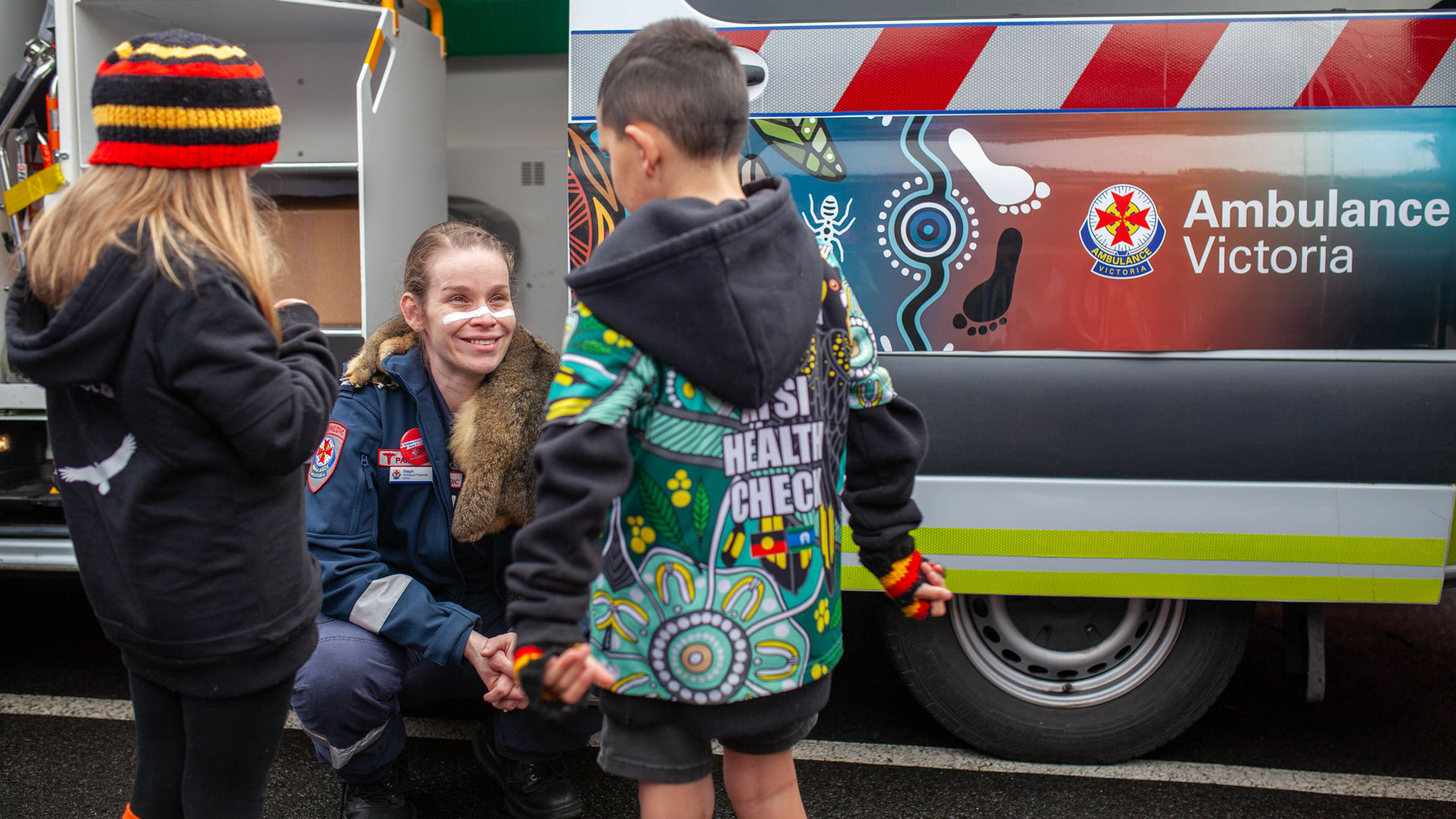 A paramedic crouches in front of an ambulance to talk to two children