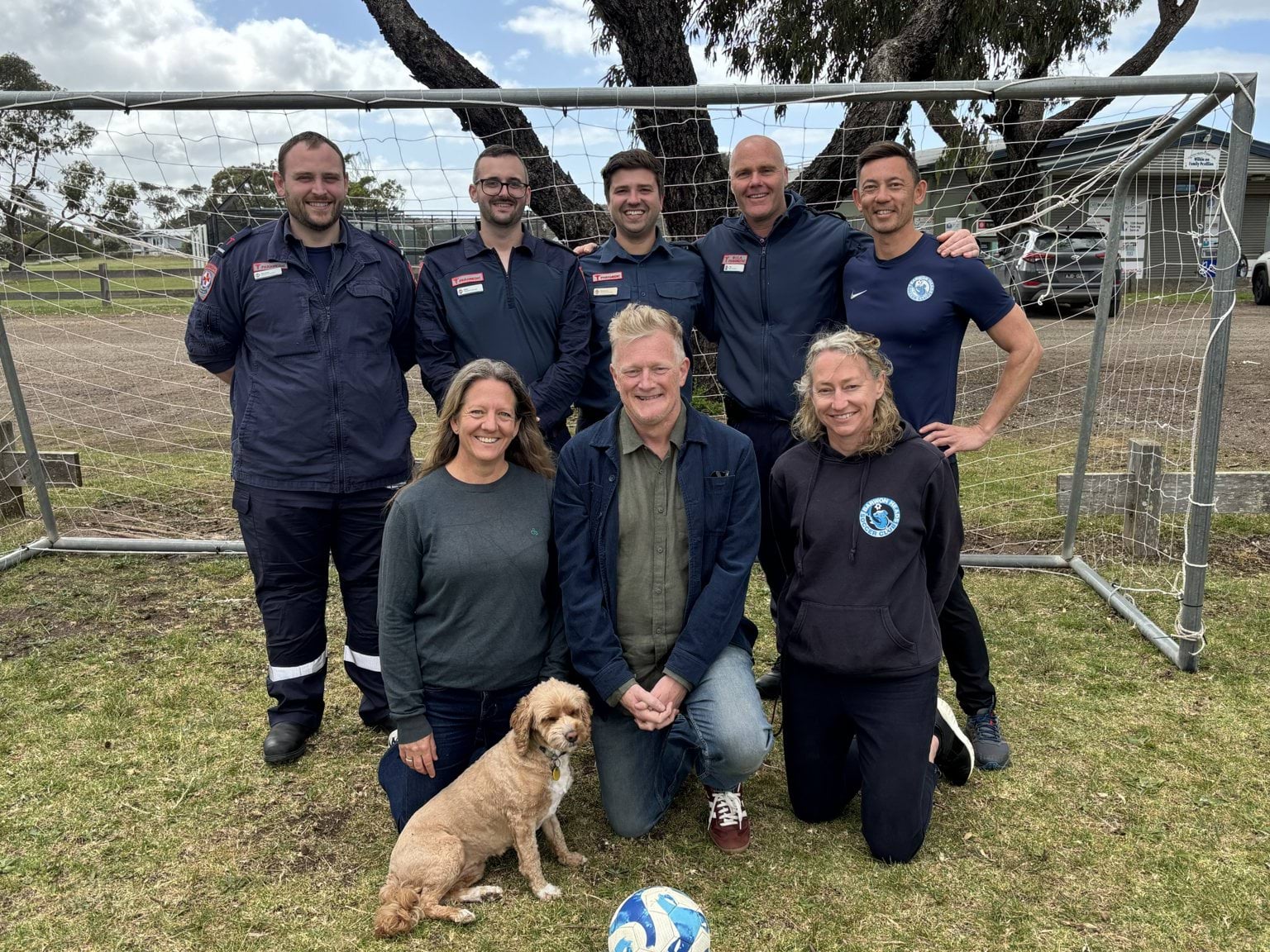 A group of four paramedics and four others in a soccer goal with a dog and soccer ball.
