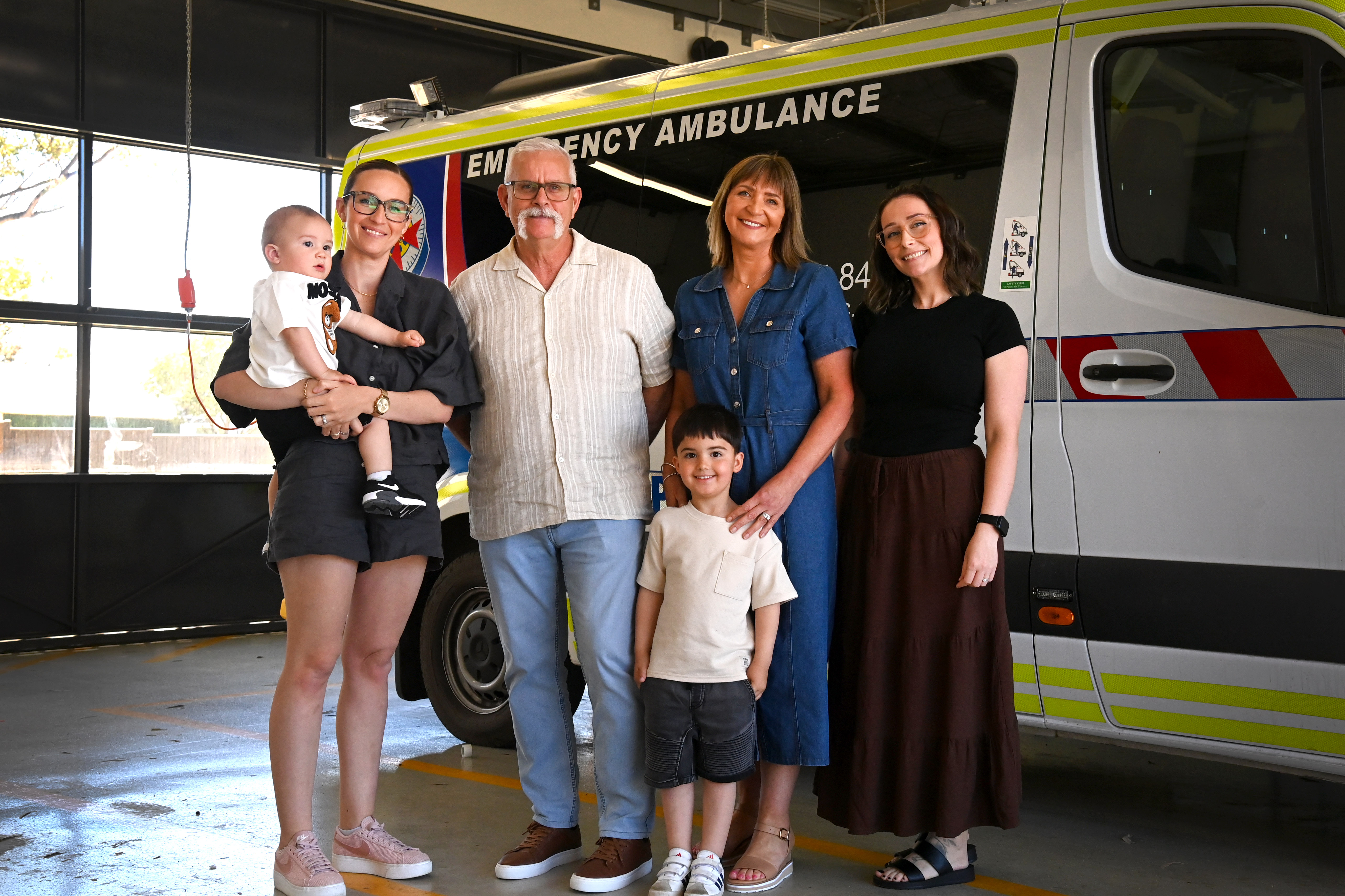 A man, three women and two young kids smile for the camera in front of an ambulance.