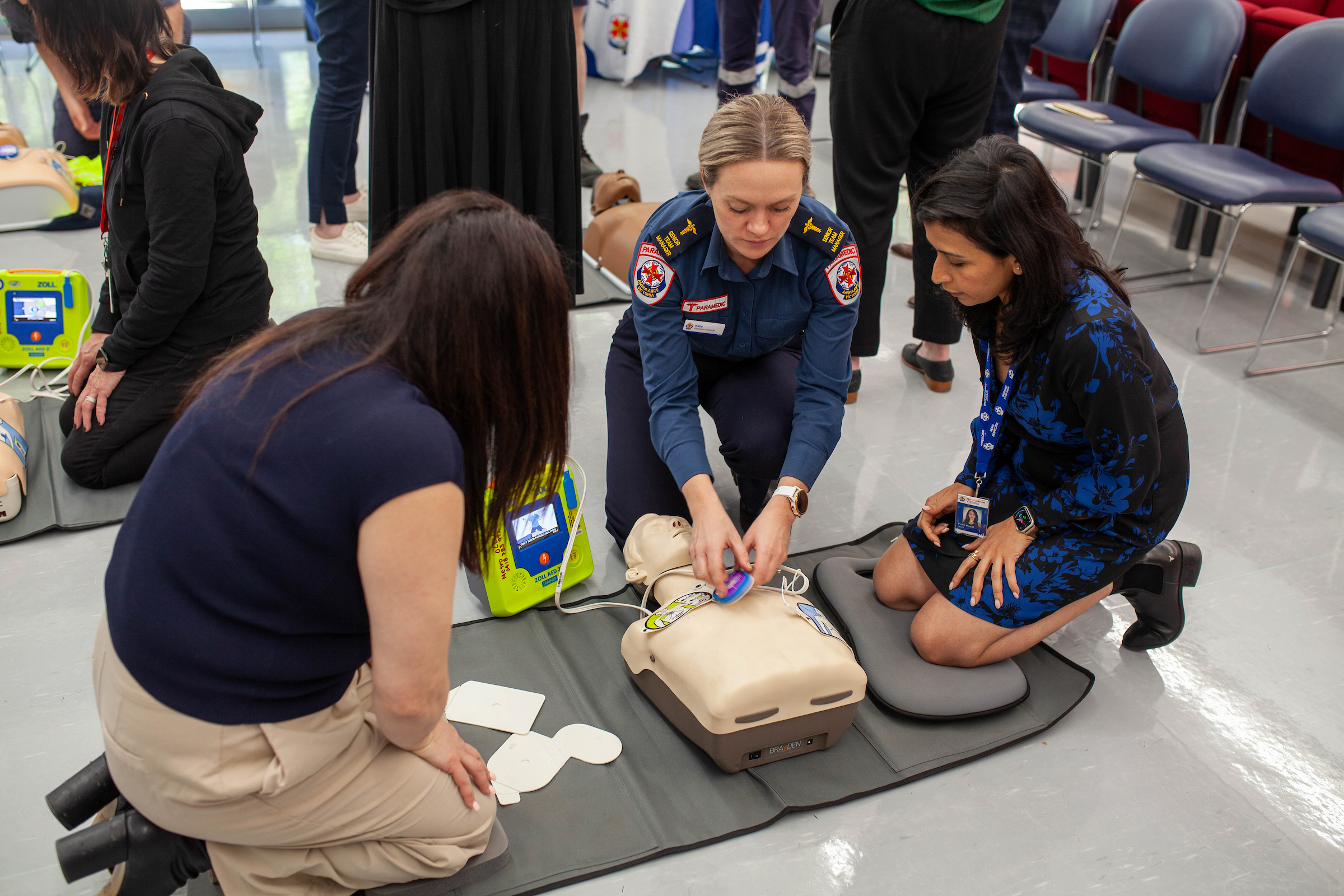 A paramedic teaches two women in business clothes how to apply a defibrillator.