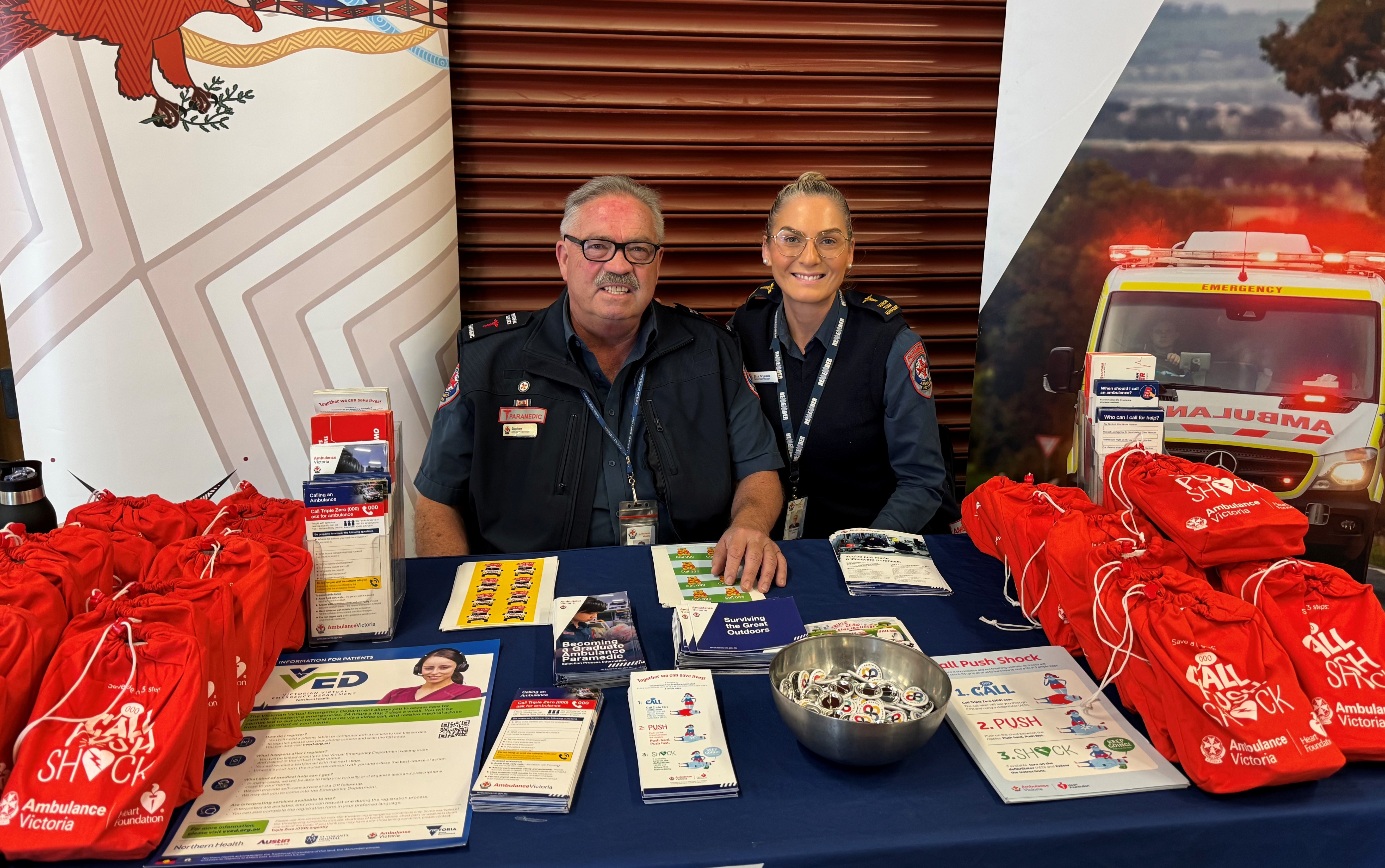 A male paramedic and a female paramedic sitting behind a table displaying brochures and red bags.