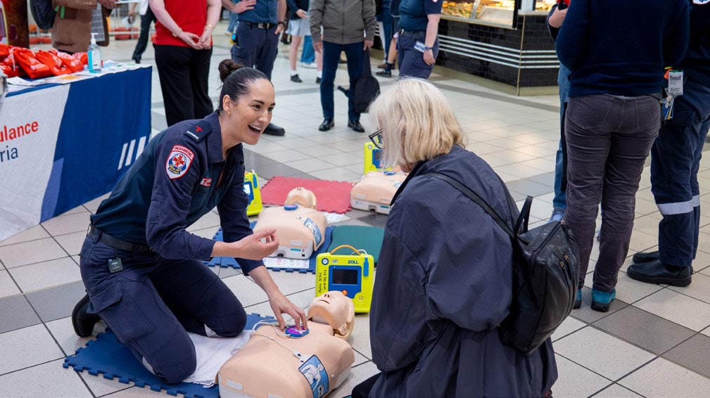 A paramedic at a train station uses a manikin to teach a woman CPR